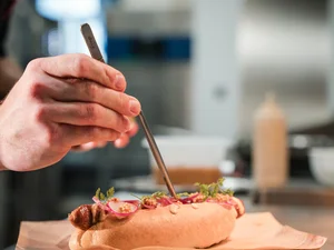 Hand, die mit Pinzette Zutaten auf einem Hotdog arrangiert
