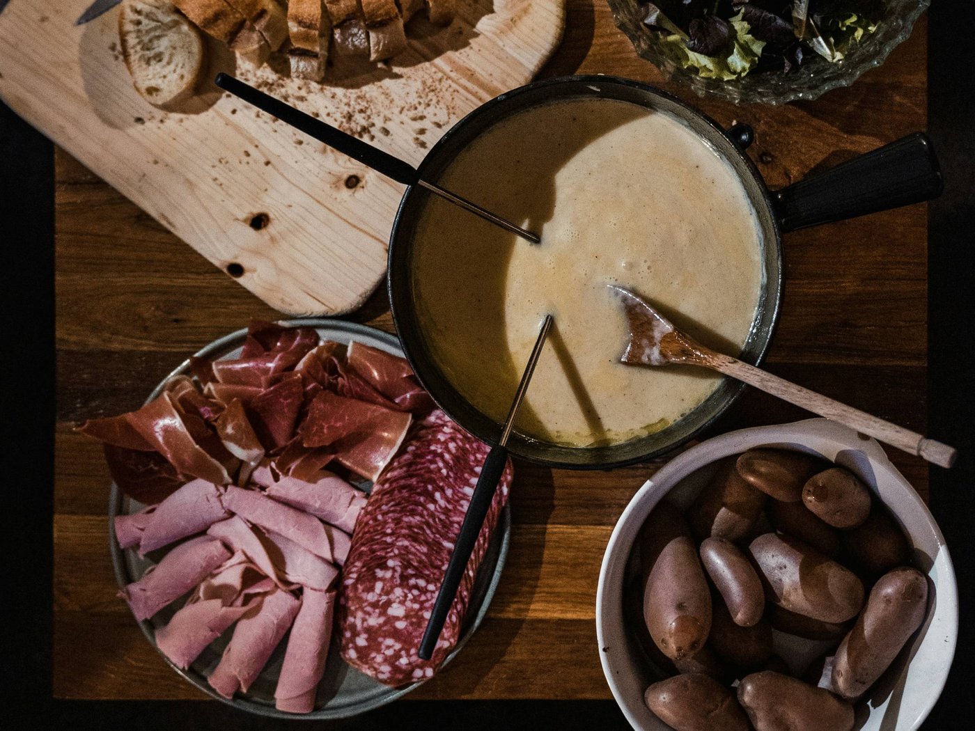 Fondue-Set mit Käse, Brot, Salat, Kartoffeln und Aufschnitt auf Holztisch