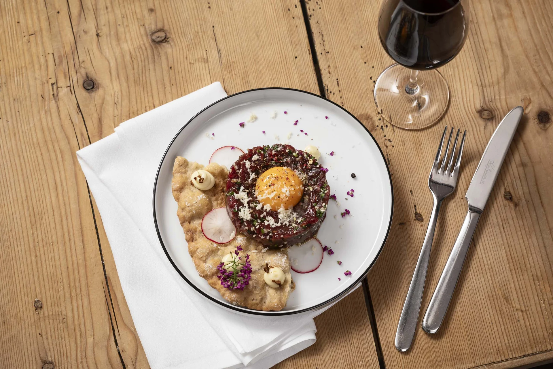Bayerisches Restaurant nahe Augsburg gesucht? Tatar mit rohem Eigelb, Radieschenscheiben, Cracker und Glas Rotwein auf Holztisch