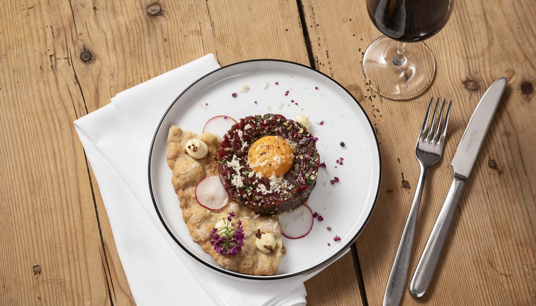 Tatar mit rohem Eigelb, Radieschenscheiben, Cracker und Glas Rotwein auf Holztisch