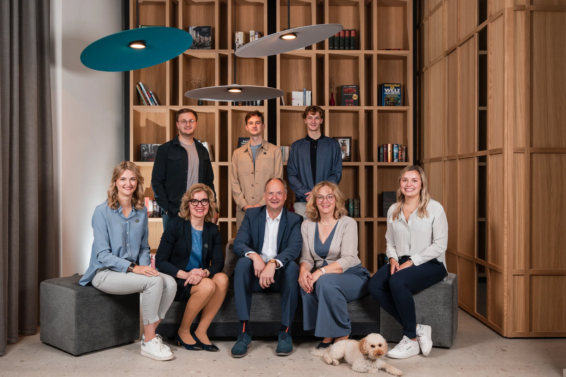 Gruppenbild von acht Menschen und einem kleinen Hund in einem modernen Raum mit Bücherregal