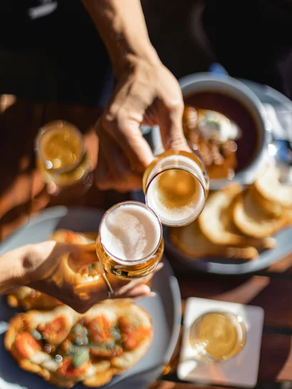 Zwei Hände stoßen mit Biergläsern an, Essen auf dem Tisch darunter