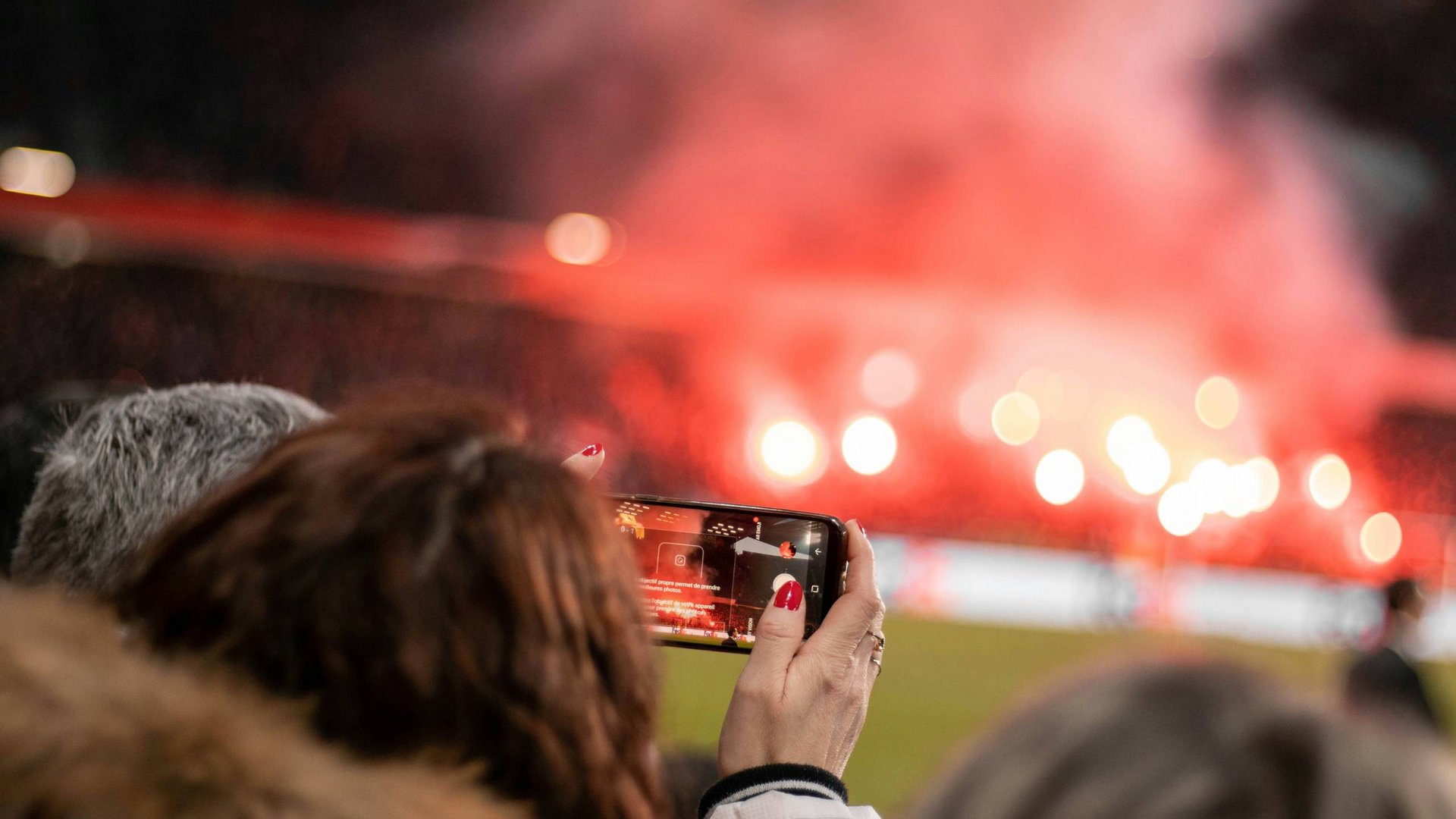 Person filmt rot leuchtendes Feuerwerk auf einem Sportplatz mit dem Smartphone