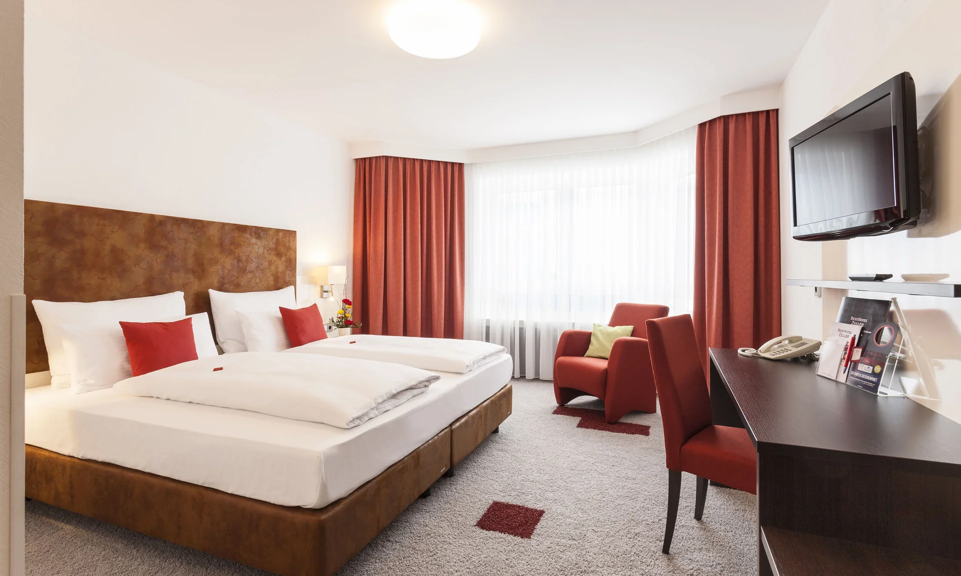 Helles Hotelzimmer mit Doppelbett, roten Vorhängen und Sesseln