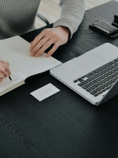 Person schreibt in Notizbuch neben offenem Laptop und Kaffeetasse auf schwarzem Tisch
