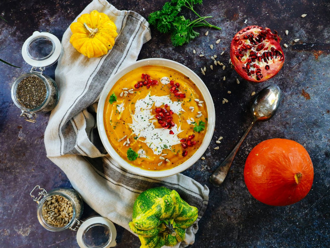 Schüssel Kürbissuppe mit Sahne, Granatapfelkernen und Kräutern umgeben von Kürbissen und Samen