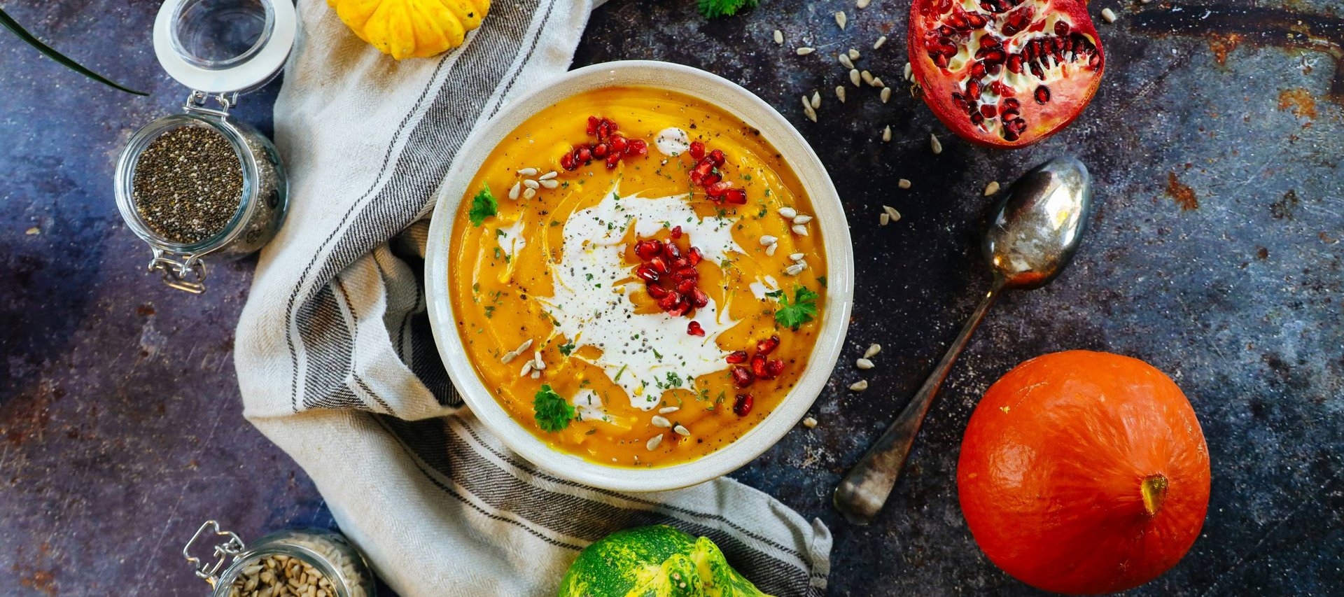 Schüssel Kürbissuppe mit Sahne, Granatapfelkernen und Kräutern umgeben von Kürbissen und Samen