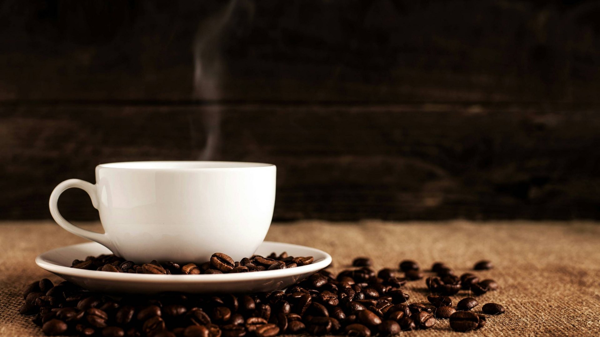 Ihr Start in den Tag: Frühstück in Königsbrunn Weiße Tasse mit dampfendem Kaffee umgeben von Kaffeebohnen auf Leinwand