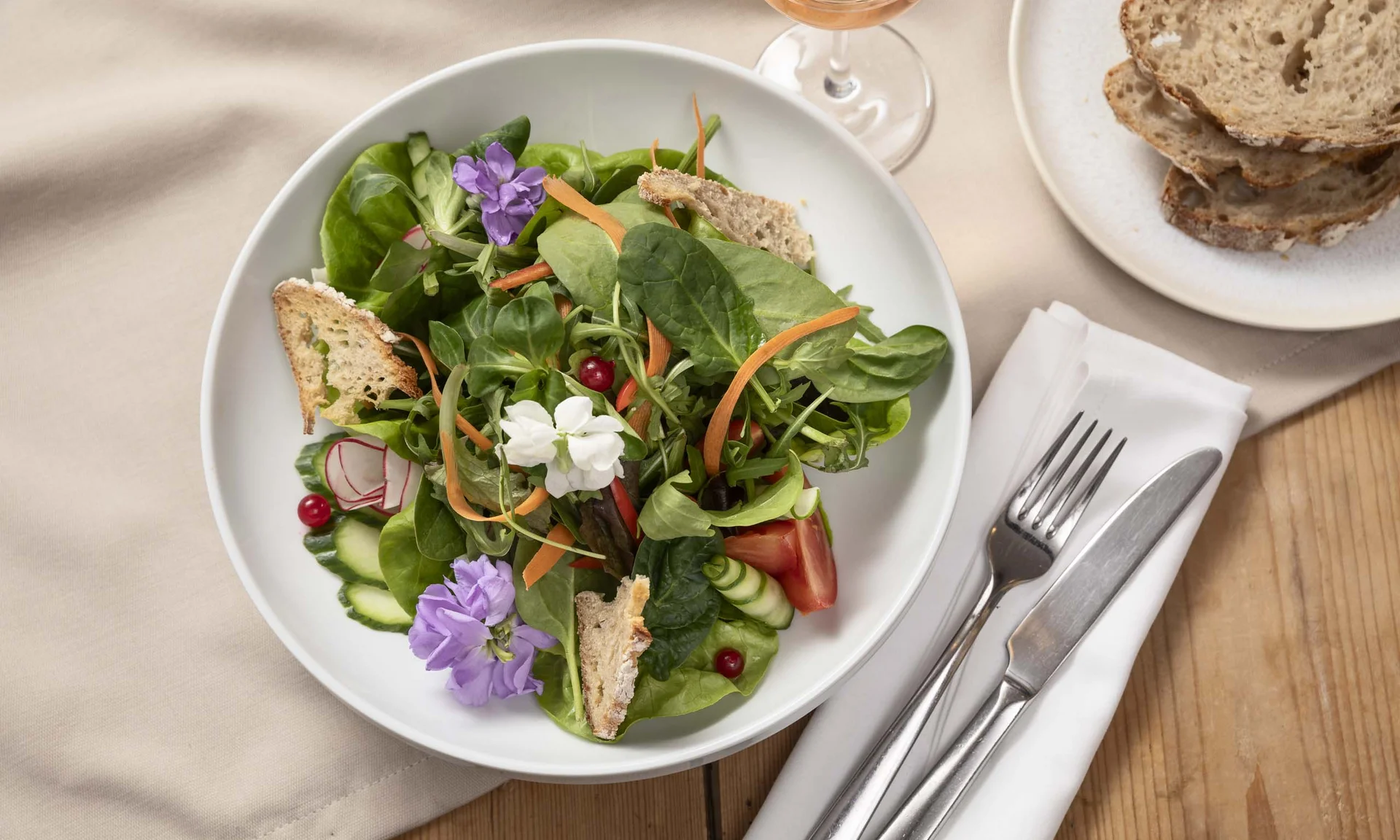 Salat mit essbaren Blumen, Brot und Besteck auf Holztisch