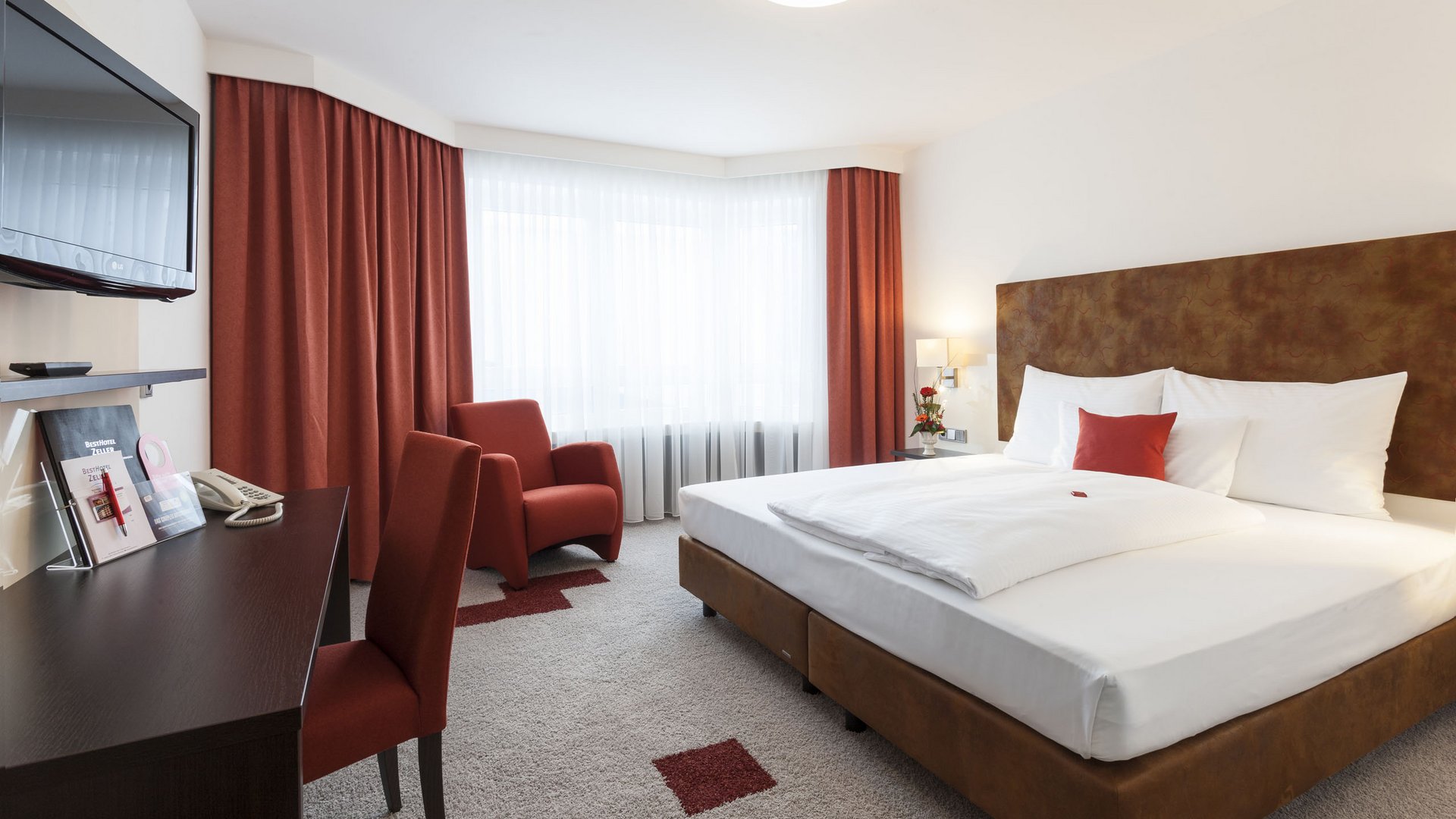 Modernes Hotelzimmer mit großem Bett, rotem Sessel, Schreibtisch und Fernseher