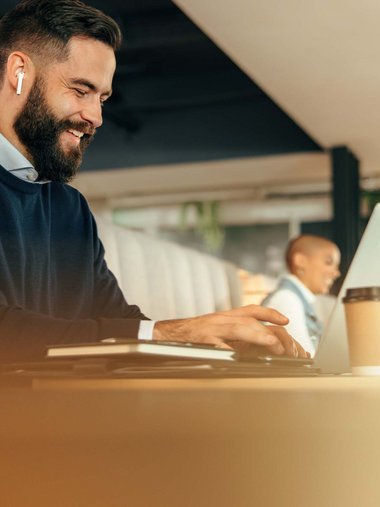 Bearded man working on laptop wearing wireless earbuds in modern office