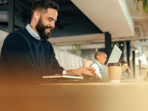 Mann mit Bart arbeitet mit Laptop und trägt kabellose Ohrhörer in modernem Büro