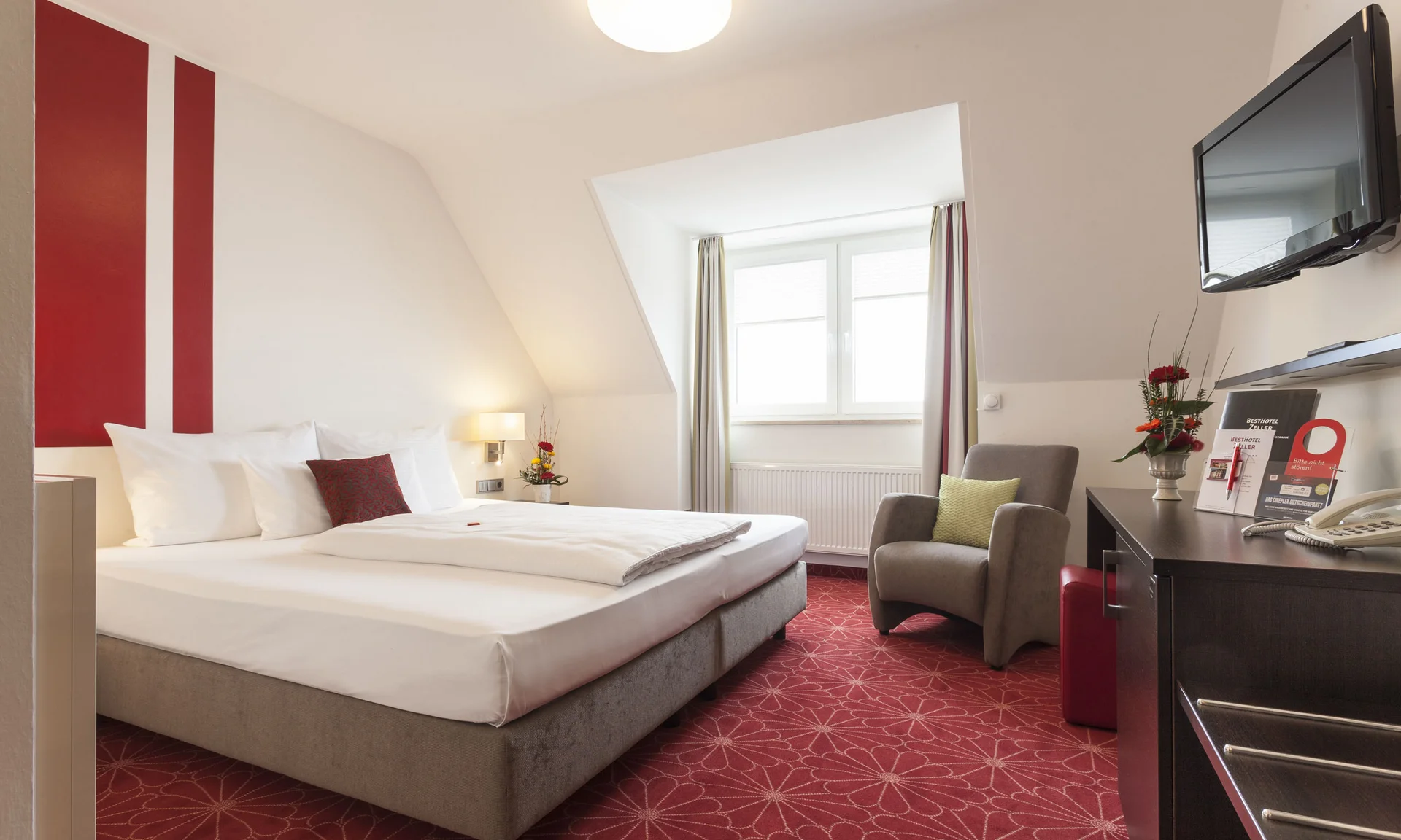 Hotelzimmer mit Doppelbett, Sesseln, TV und roten Teppichboden