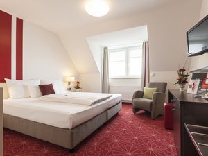 Hotelzimmer mit Doppelbett, Sesseln, TV und roten Teppichboden