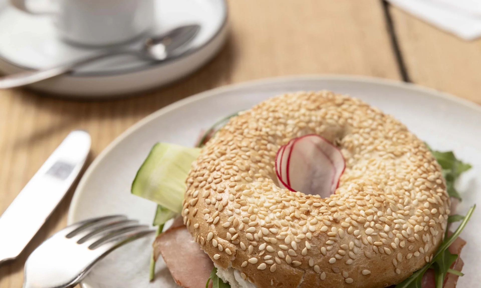 Sesam-Bagel mit Schinken, Rucola und Gurke auf weißem Teller mit Kaffee im Hintergrund