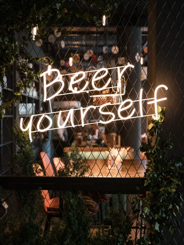 Neonlicht mit dem Text Beer yourself an einem Zaun vor einem Café