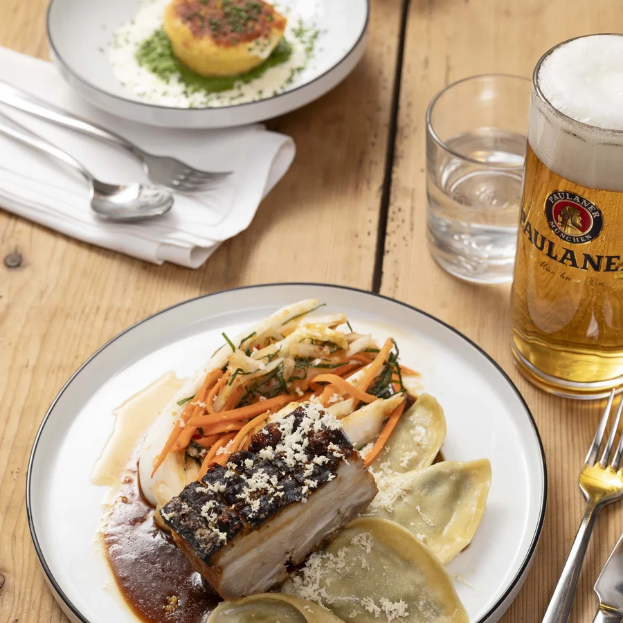 Bayerisches Restaurant nahe Augsburg gesucht? Teller mit Schweinebauch, Gemüse, Ravioli und Glas Bier auf Holz-Tisch