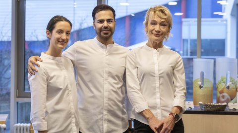 Drei freundlich lächelnde Mitarbeiter in weißer Arbeitskleidung vor Getränkespendern