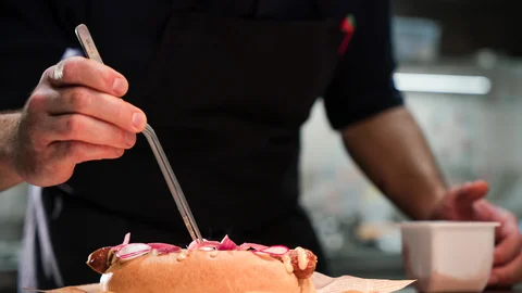 Koch dekoriert Gourmet-Hotdog mit Zange in professioneller Küche
