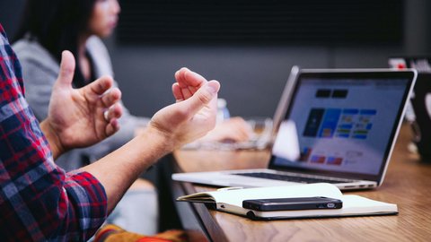 Person gestikuliert bei einer Besprechung mit Laptop und Notizbuch auf dem Tisch