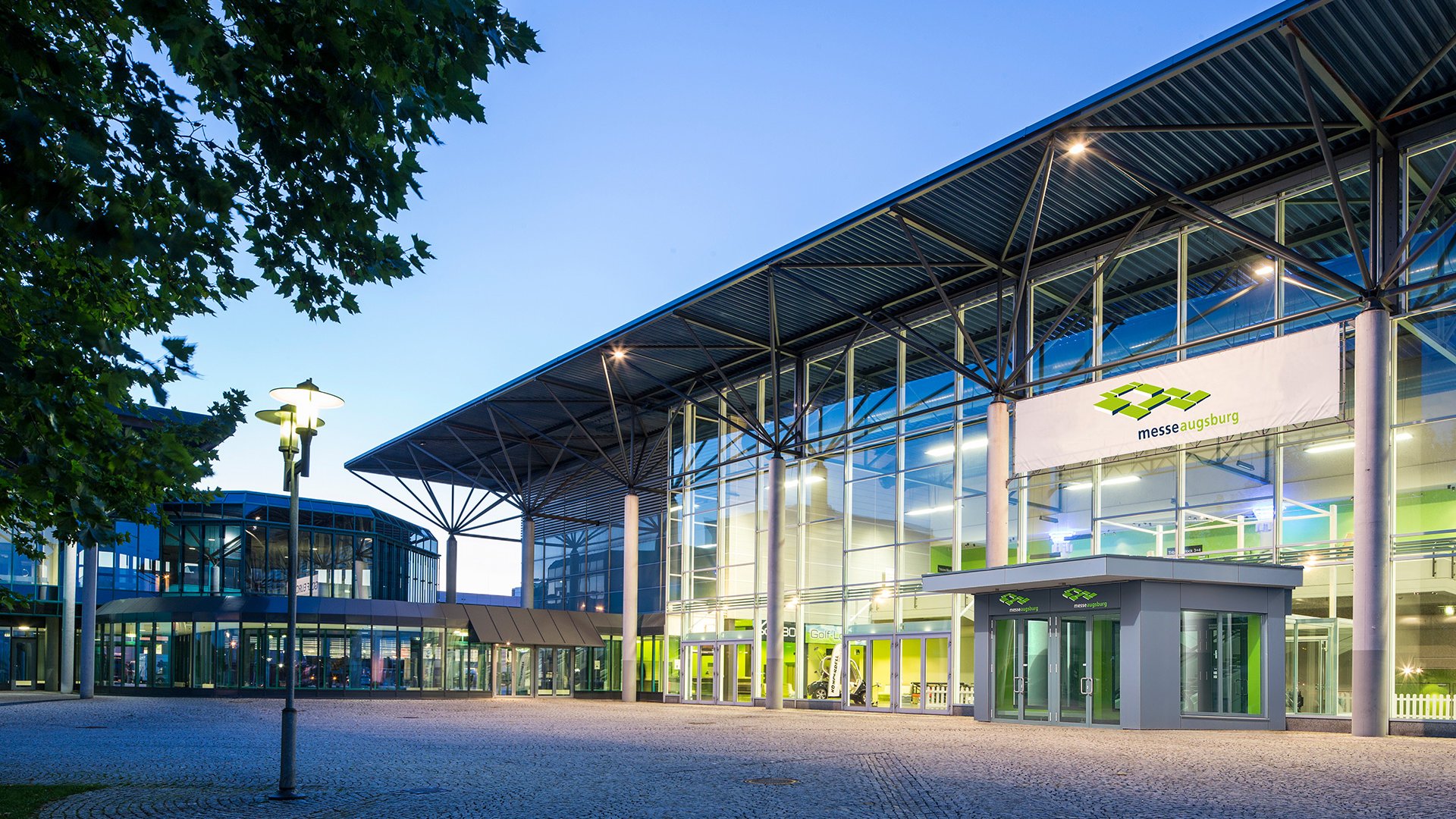 Beleuchtetes Messezentrum Augsburg bei Abenddämmerung mit Glasfassade und Bäumen