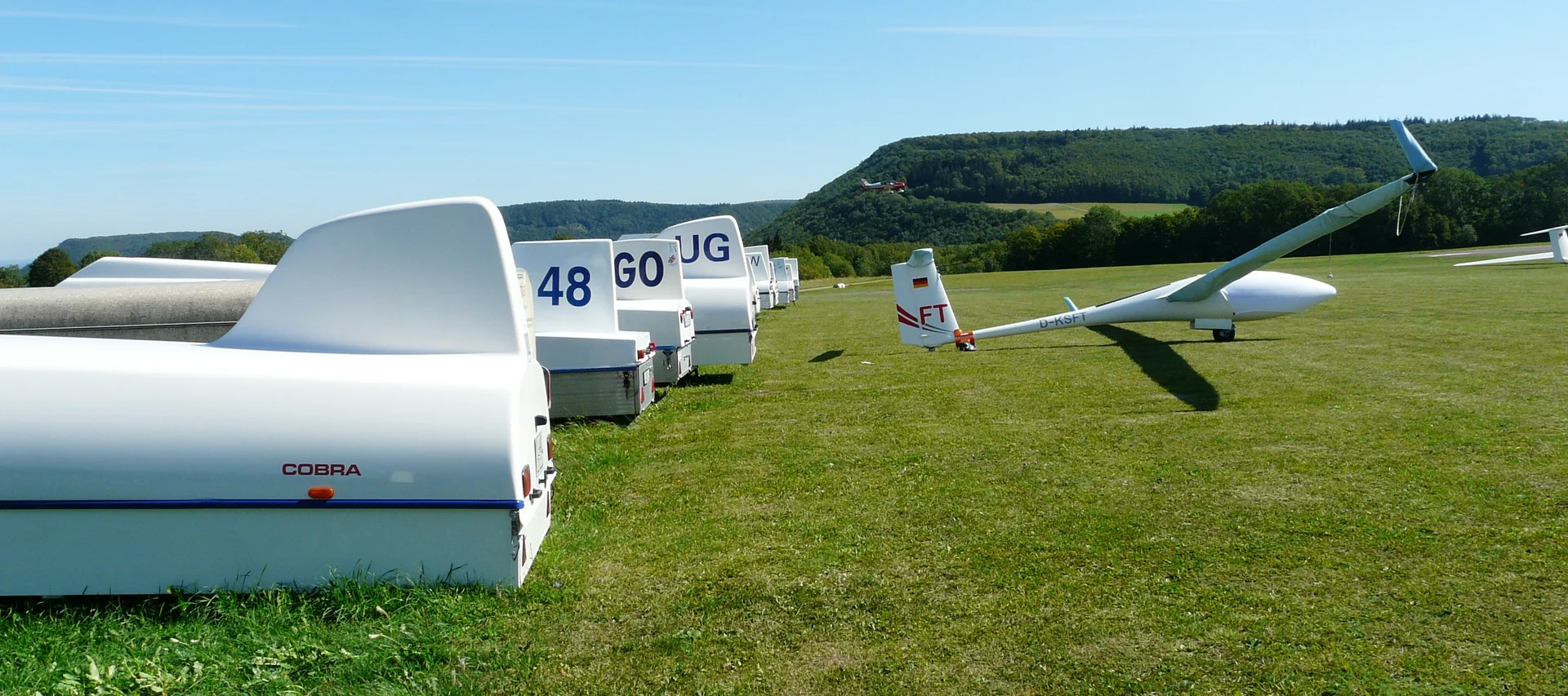 Row of gliders on grassy field with forest hills and blue sky